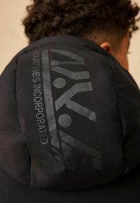 Čierna puffa bunda s hladkou, matnou textúrou a výrazným grafickým dizajnom na kapucni s textom "INDUSTRIES INCORPORATED".