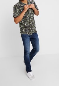 Kortärmad skjorta med ett mörkt mönster av geometriska former i grön, gul och vit; kombinerad med åtsittande blå jeans och vita sneakers.