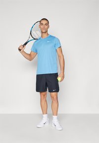 Hele sinine lühikeste varrukatega sportlik särk ja tumesinised lühikesed püksid, millel on valge Nike logo, hoides tennisereketit ja rohelist tennisepalli.