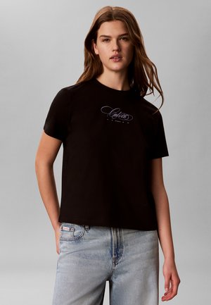 Ung kvinde iført en sort Calvin Klein t-shirt med korte ærmer og lyseblå jeans, stående foran en ensfarvet grå baggrund.
