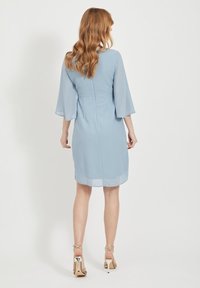 Žena s vlnitými rudými vlasy, oblečená v světlemodrých šatech ke kolenům s tříčtvrtečními rukávy a zlatými vysokými sandály, stojící zády.