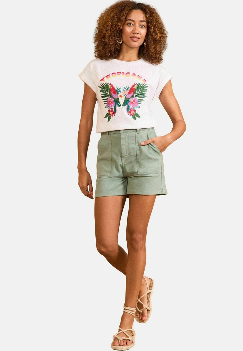 Chemise blanche à manches courtes avec imprimé tropical ; shorts taille haute verts ; sandales beiges. Le modèle se tient avec une main dans sa poche.