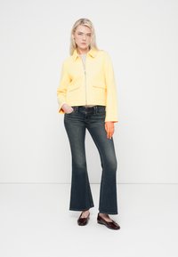 Κίτρινο σακάκι με γιακά και μπροστινές τσέπες, συνδυασμένο με σκουρομπλε τζιν καμπάνα. Το μοντέλο φοράει σκούρα μοκασίνια και στέκεται μπροστά σε λευκό φόντο.