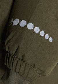 Rukáv olivovo zelenej bundy má štruktúru herringbone, elastický manžet a strieborné kruhové logo s tlačeným vzorom bodiek pozdĺž rukáva.