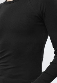 Κοντινό πλάνο ενός ατόμου που φοράει εφαρμοστή μαύρη μπλούζα με μακριά μανίκια και στρογγυλή λαιμόκοψη σε απλό ανοιχτό φόντο.