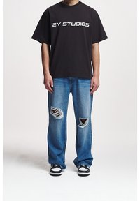Person wearing black "2Y STUDIOS" t-shirt, ripped blue jeans with holes on knees, and black-and-white sneakers standing against a plain background.