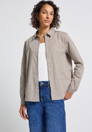 Kvinna klädd i en beige och vit randig skjorta med knappar över en vit topp, ihop med blå jeans, stående mot en enkel vit bakgrund.