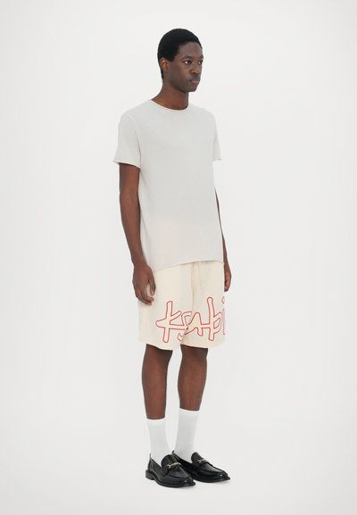 Camiseta de manga corta gris claro, pantalones cortos beiges con impresión roja de 'ksabi', calcetines tobilleros blancos y mocasines negros. Diseño simple y moderno.