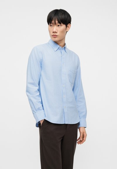 Junger Mann, der ein hellblaues Hemd mit Brusttasche und dunkle Hose trägt, steht mit einer Hand in der Tasche und schaut seitlich.