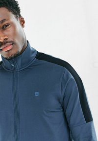 Granatowa sportowa kurtka z wysokim kołnierzem, zamkiem błyskawicznym i czarnymi akcentami na ramionach. Na piersi małe niebieskie logo, gładka faktura.