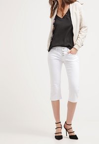 White cropped jeans with five-button closure, paired with a black v-neck top and a light cream bomber jacket with floral accents. Black high-heeled shoes.