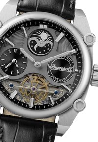 Montre automatique en argent avec un cadran gris texturé, des sous-cadrans, des engrenages visibles, un bracelet en cuir noir et un boîtier en acier inoxydable.