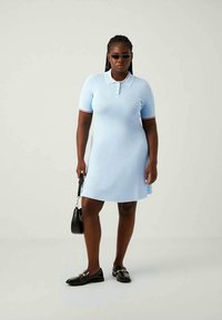 Mujer vestida con un vestido polo de manga corta azul claro, mocasines negros, gafas de sol y sosteniendo un pequeño bolso negro, de pie frente a un fondo liso.