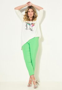 Top bianco a maniche lunghe con stampa di cocktail; abbinato a pantaloni fitted verdi brillanti e sandali argentati con cinturini, creando un insieme vivace.