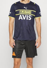 Tummansininen urheilupaita keltaisin koristein, jossa on "FENERBAHÇE" ja "AVIS" -brändäys. Yhdistetty mustiin shortseihin ja kolmionmuotoiseen oranssiin logoon.