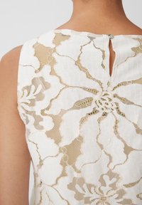 Vit spetsöverdel med blommönster, texturerad insättning och en nyckelhålsdetalj bak som är säkrad med en liten rund knapp.