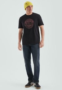 Czarna koszulka T-shirt z okrągłym burgundowym napisem, w połączeniu z ciemnoniebieskimi dżinsami i czarnymi sneakersami z brązowymi podeszwami; model nosi żółtą czapkę.