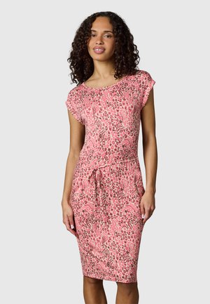 Kobieta o kręconych włosach, ubrana w różowo-brązową sukienkę z nadrukiem zwierzęcym do kolan, z rękawami typu cap i paskiem w talii, stojąca na jednolitym tle.