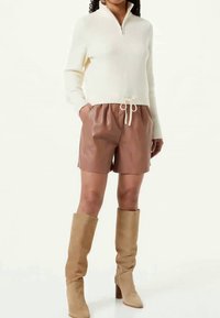 Γυναίκα με κρεμ ριμπ ζακέτα με φερμουάρ, καφέ δερμάτινα σορτς με ρυθμιζόμενο κορδόνι και καμβά μπότες μέχρι το γόνατο σε απόχρωση ταμπά, που στέκεται όρθια.