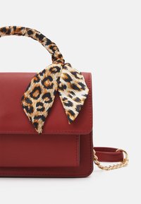 Borsa a tracolla in pelle rossa con una sciarpa con stampa leopardata legata al manico e dettagli in oro. Design semplice e rettangolare con chiusura a pattina.