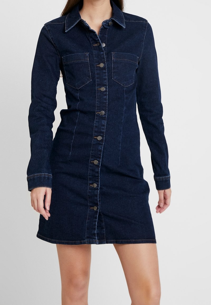Ciemnogranatowa sukienka koszulowa z długimi rękawami, zapięciem na guziki, dwoma kieszeniami na piersiach i dopasowanym krojem, ozdobiona jasnymi przeszyciami.