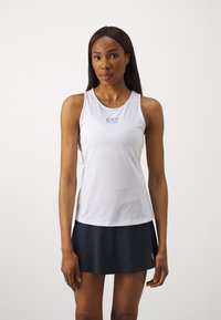 Top sportivo bianco senza maniche con logo sul petto, abbinato a una gonna blu scuro. Tessuto morbido e design senza maniche per l'abbigliamento attivo.