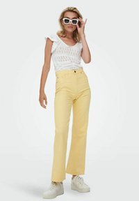 Top blanco de punto con mangas cortas con volantes, combinado con pantalones amarillos de talle alto y pierna recta, y zapatillas de plataforma blancas.