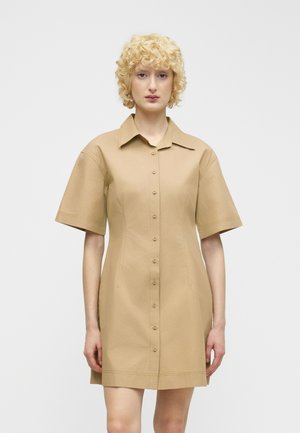Femme aux cheveux blonds bouclés portant une robe beige boutonnée à manches courtes, arrivant au genou, debout devant un fond blanc uni.