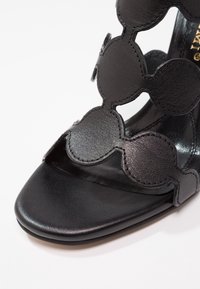 Sandale en cuir noir avec un design en découpes festonnées, une texture lisse et une semelle plate avec un bout arrondi.