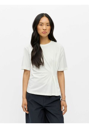 Femme aux longs cheveux noirs porte un haut blanc à manches courtes avec des plis latéraux et un pantalon noir à jambes larges sur fond uni.