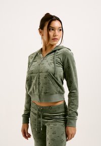 Green velour zip-up hoodie with drawstrings, featuring a front pocket and subtle brand logo patterns. Paired with matching jogger pants.