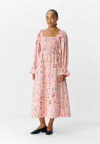 Vestido largo rosa de manga larga con escote cuadrado, que presenta un cuerpo fruncido y un estampado de varias formas y colores, combinado con zapatos negros.