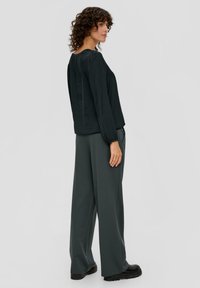 Schwarze Bluse mit langen Ärmeln, lockerer Passform, Rundhalsausschnitt und elastischen Manschetten, kombiniert mit dunkelgrauen weiten Hosen und schwarzen klobigen Schuhen.
