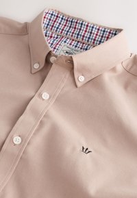 Camicia beige con bottoni e collo alla coreana con bottoni, bottoni bianchi, fodera interna del colletto a quadretti rossi e blu e piccolo logo corona nero sul petto.