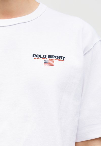 Λευκό T-shirt με το κείμενο "Polo Sport Ralph Lauren" και μια μικρή αμερικανική σημαία κεντημένη στο αριστερό μέρος του στήθους.