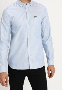 Camisa azul claro de algodón con botones, manga larga, bolsillo en el pecho y un pequeño logo amarillo en el bolsillo.