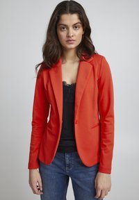 Rode blazer, gestructureerd ontwerp, sluiting met één knop, twee voorzakken, gedragen over een zwarte kanttop, in combinatie met blauwe jeans.