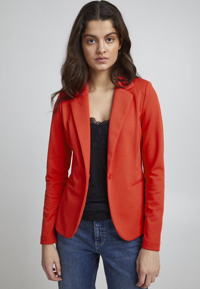 Rode blazer, gestructureerd ontwerp, sluiting met één knop, twee voorzakken, gedragen over een zwarte kanttop, in combinatie met blauwe jeans.