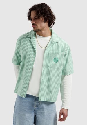 Man with curly hair wearing mint green striped short-sleeve shirt over white long sleeves and light blue jeans, looking left.