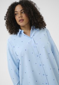 Chemise bleu clair à boutons avec col, ornée de petits motifs de cœurs bleus, manches longues et coupe décontractée. Texture de tissu lisse.