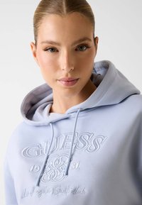 Felpa con cappuccio azzurro chiaro realizzata in tessuto liscio. Presenta un logo "GUESS" ricamato con dettagli decorativi. I laccetti sono visibili all'altezza del collo.