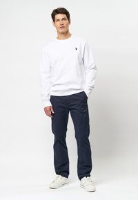 Witte sweatshirt met lange mouwen en ribgebreide manchetten, versierd met een klein logo, gecombineerd met donkerblauwe chinos en witte sneakers, staande voor een neutrale achtergrond.