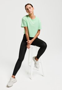 T-shirt court vert clair avec logo, associé à un legging noir et des baskets de couleur claire. Modèle assis sur un tabouret blanc.