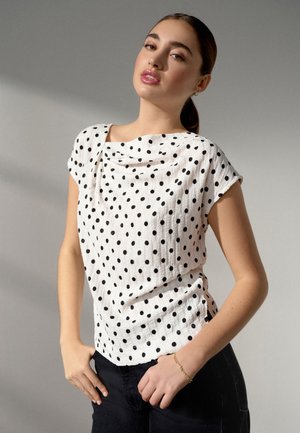 Giovane donna con i capelli raccolti indossa una camicetta bianca a maniche corte con pois neri e pantaloni neri, in piedi davanti a uno sfondo semplice.