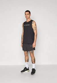 Muškarac s tetovažama nosi crni bezrukavni top HYROX, crne kratke hlače, bijele Puma čarape i crne tenisice dok stoji protiv prazne pozadine.