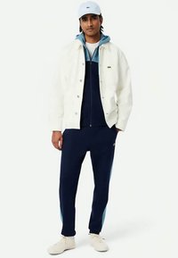 Άνδρας φορώντας λευκό μπουφάν, μπλε και ανοιχτό μπλε φούτερ με φερμουάρ, ναυτικό μπλε αθλητικά παντελόνια, λευκό καπέλο και λευκά αθλητικά παπούτσια, στέκεται με το ένα χέρι στην τσέπη.