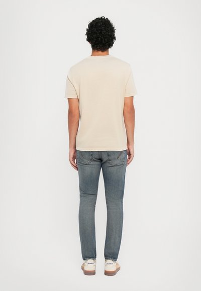 Beige bomulds t-shirt med korte ærmer og rund halsudskæring, parret med slim-fit blå jeans og hvide sneakers med brune såler.