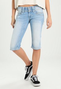 Světle modré denimové capri kalhoty s přiléhavým střihem, dvěma předními kapsami a knoflíkovým zapínáním, spojené s černými teniskami. Hladká textura.