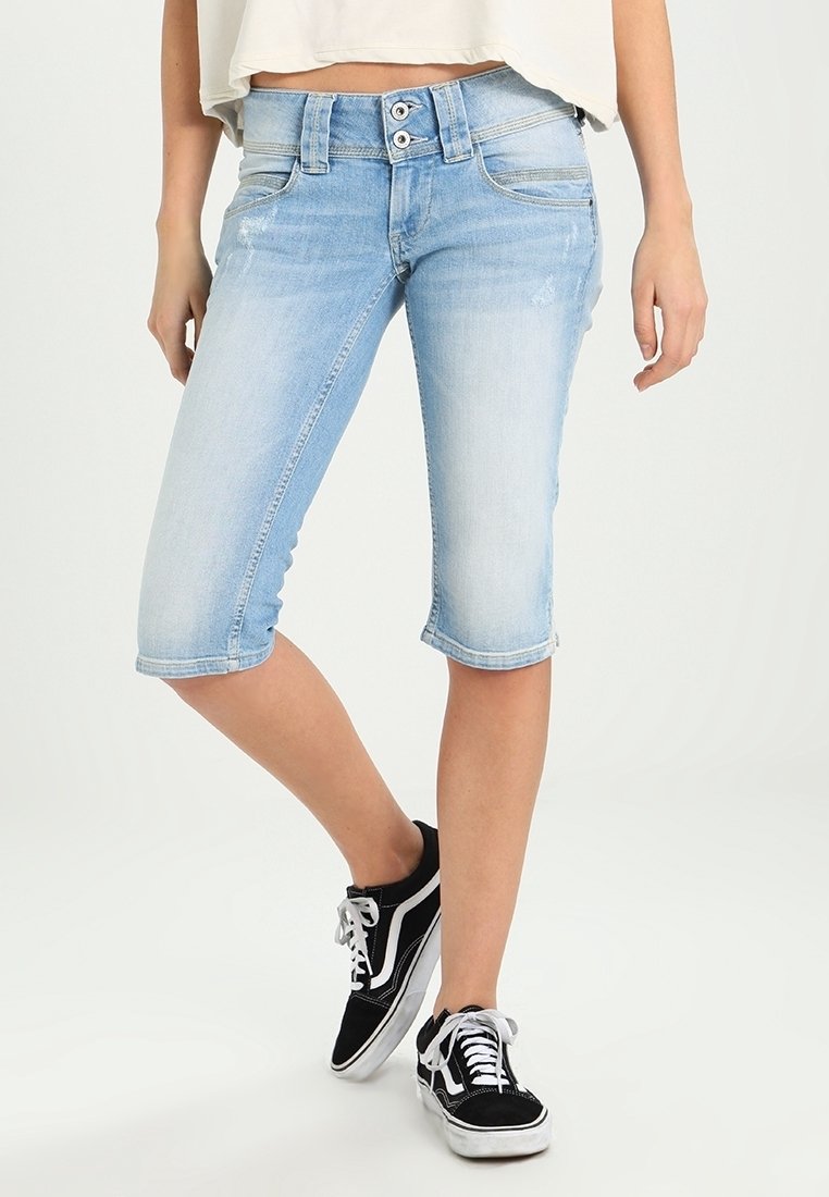 Světle modré denimové capri kalhoty s přiléhavým střihem, dvěma předními kapsami a knoflíkovým zapínáním, spojené s černými teniskami. Hladká textura.