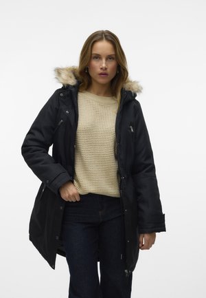Parka noire avec une capuche en fourrure, dotée de fermetures à glissière et de boutons-pression, portée par-dessus un pull beige texturé et un jean foncé.